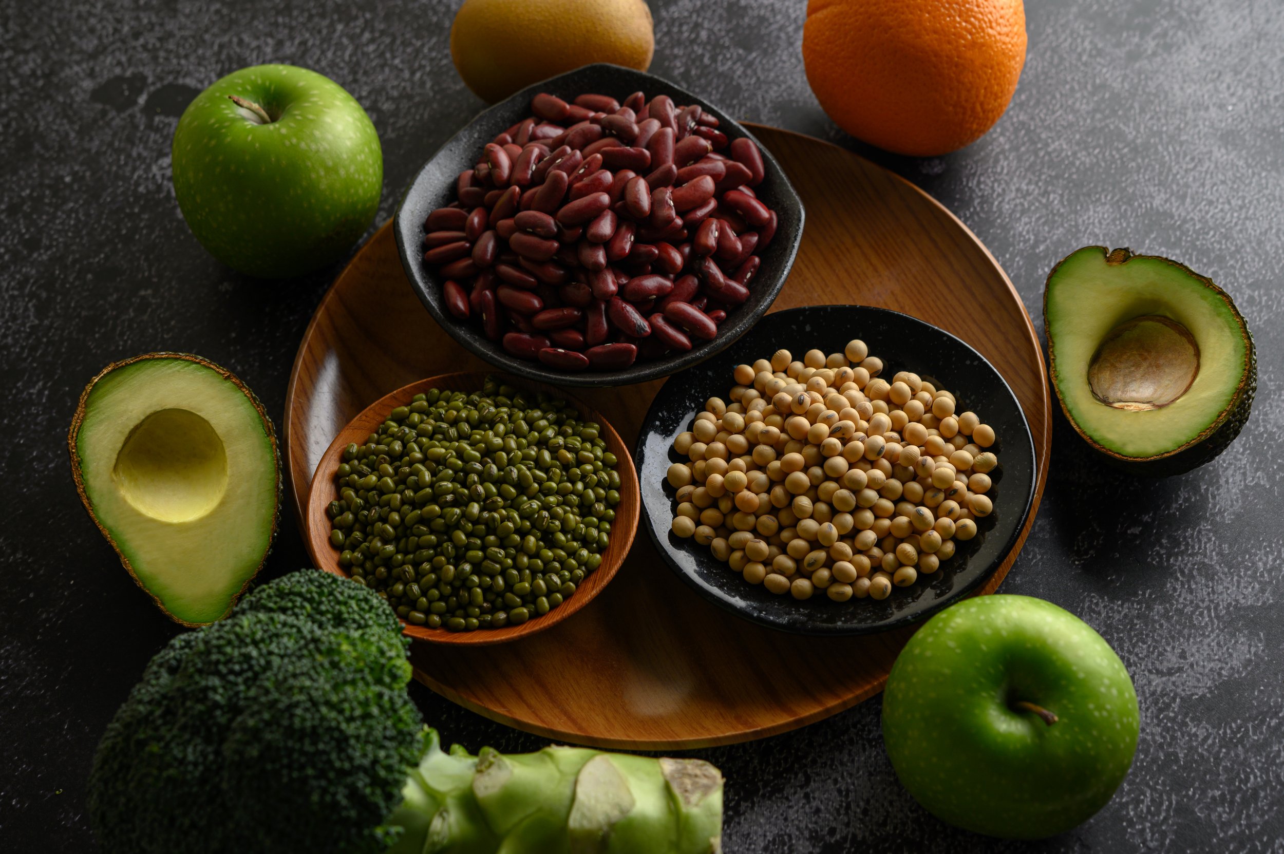 Variety of fruits and vegetables, including avocado halves, green apples, broccoli, an orange, mung beans, soybeans, and red kidney beans displayed on a dark surface.