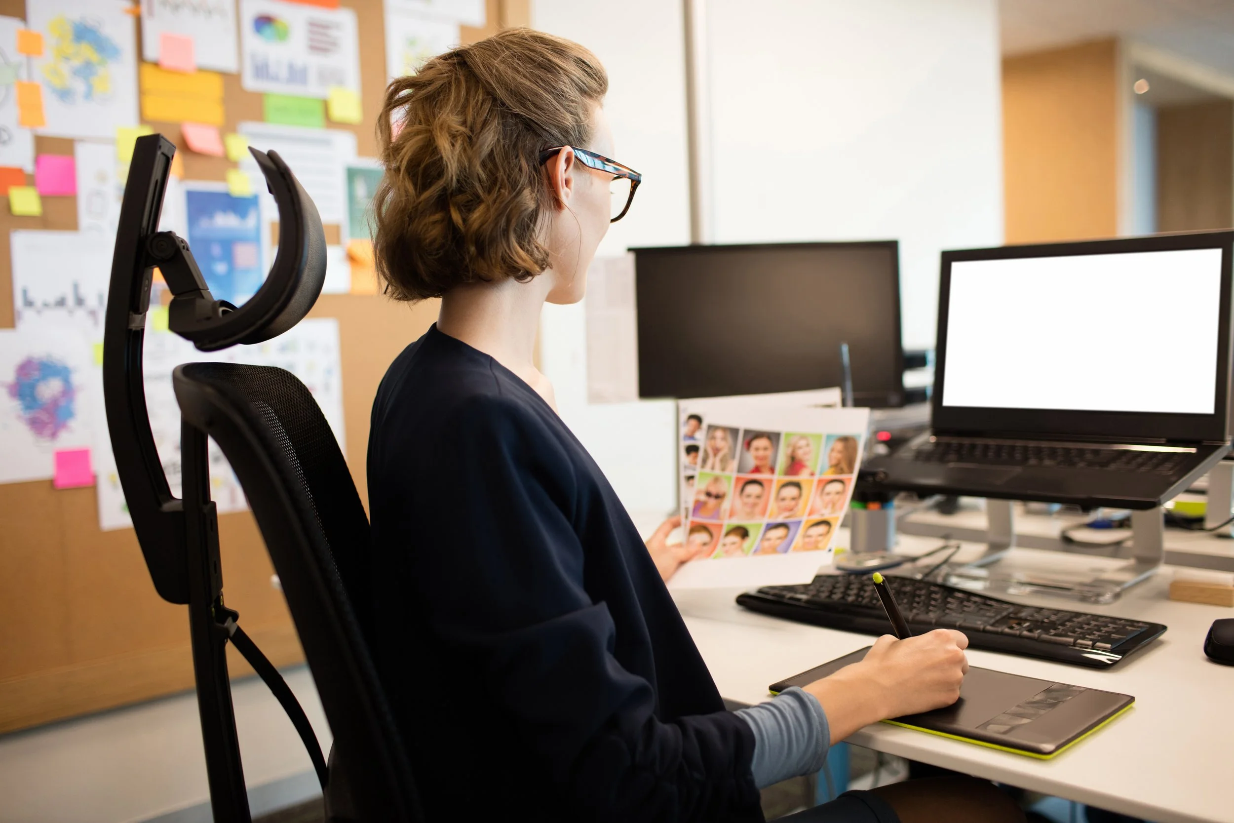Woman in office using drawing tablet in front of computer, holding sheet with headshots, cork board with sticky notes and charts on wall.